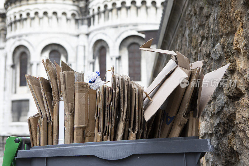 可回收的折叠纸板堆放在一个有轮子的垃圾箱里，Sacré-Cœur Basilica在远处